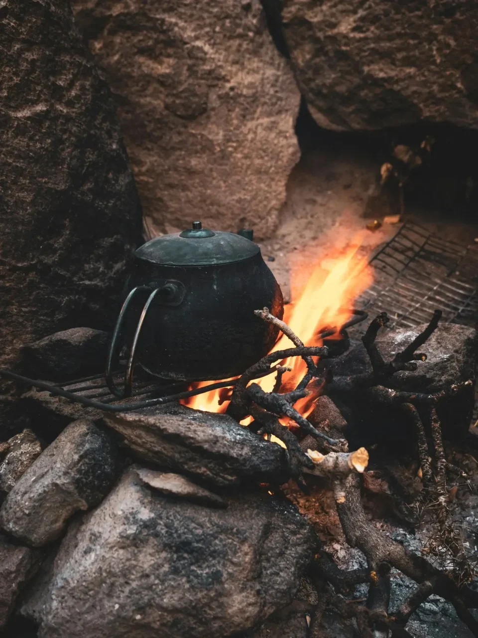 A vintage teapot sits on a campfire surrounded by rocks, evoking a rustic outdoor camping scene.